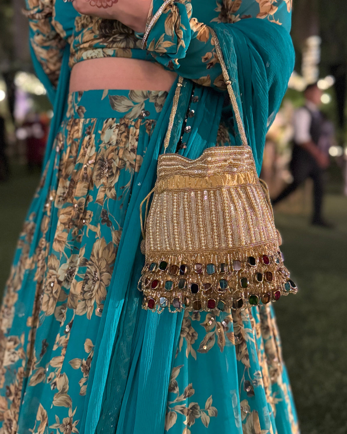 Teal embroidered traditional outfit with gold details and a handbag, blurred background